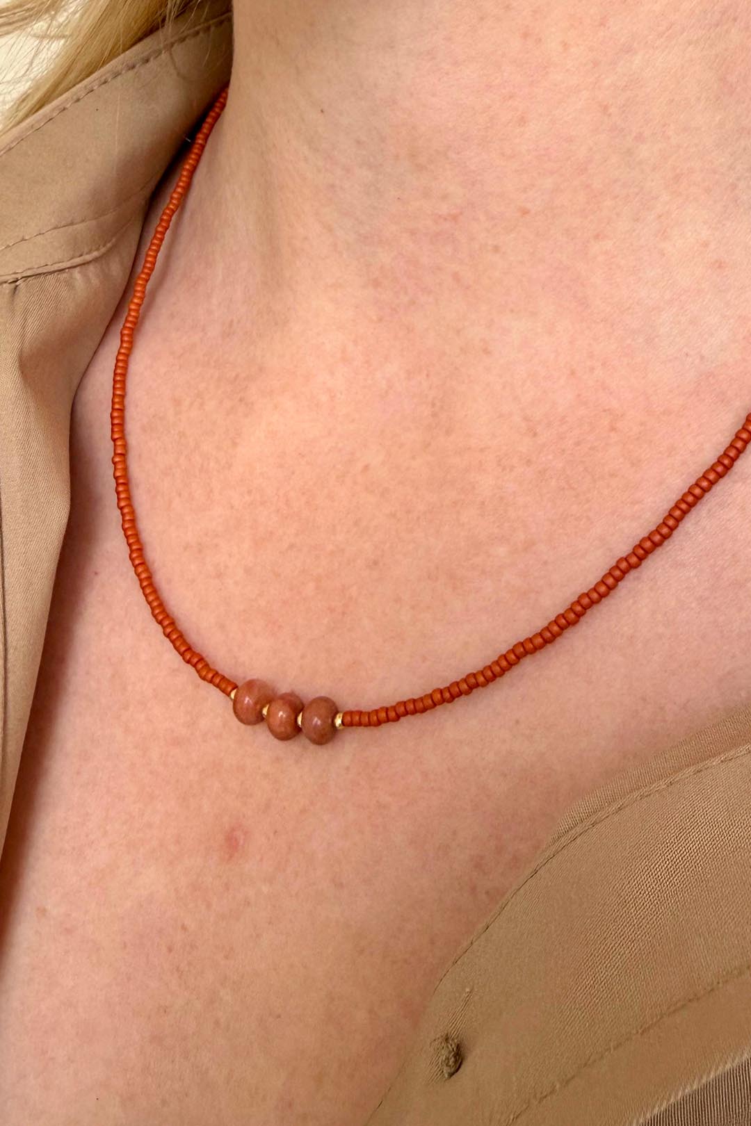 Close-up van een vrouw die een minimalistische oranje kralenketting draagt met drie centrale terracotta kralen en gouden details, gecombineerd met een beige blouse 2